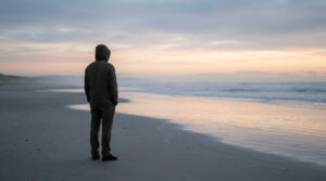 Une personne solitaire en capuche regarde l'horizon sur une plage au coucher du soleil.